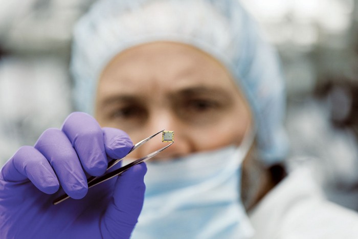 Photo of a technician in clean-room garb holding a computer chip up for inspection with tweezers. 09835-feature2-tweeze.jpg