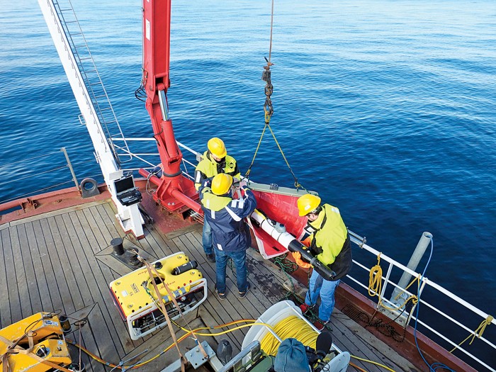 Three researchers in hard hats stand on the deck of a ship preparing to launch a torpedo-shaped underwater vehicle. 09837-polcon3-ship.jpg