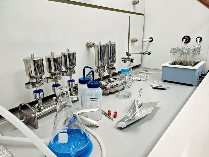 Photograph showing the benchtop inside a lab hood, with many pieces of equipment visible including metal containers, a glass flask, and tubes covered with aluminum foil.